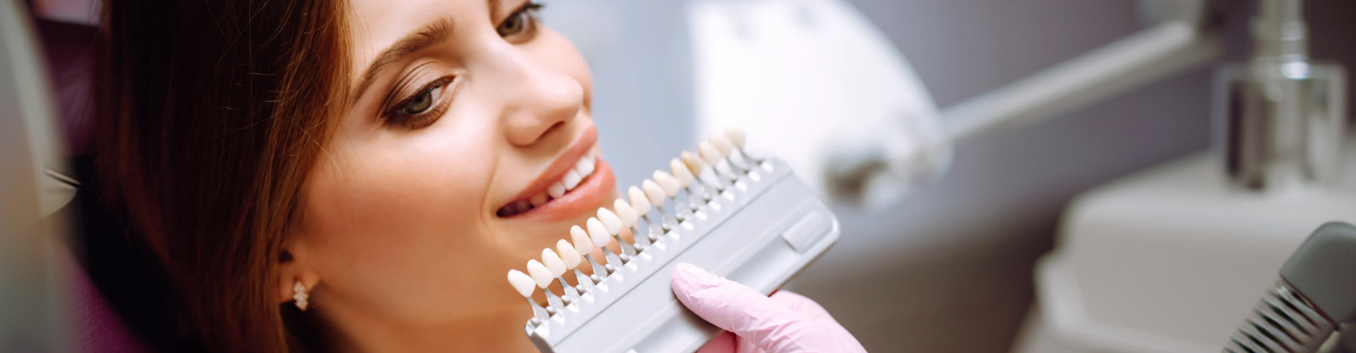 Smiling dental patient comparing her teeth to a shade guide during a consultation for porcelain veneers.